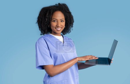 Photo for Positive black millennial lady nurse in uniform typing on computer, isolated on blue studio background. Help, exam remotely with technology, work, professional occupation, medicine and health care - Royalty Free Image