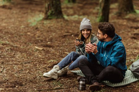 Téléchargez les photos : Joyeux jeune couple caucasien en vestes dans la forêt boire des tasses de thé de thermos, profiter des voyages, des vacances et du café chaud, en plein air. Repos, randonnée, mode de vie, aventure et tourisme - en image libre de droit