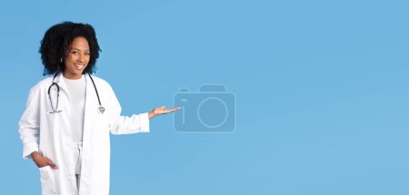 Glad black millennial lady doctor in white coat hold empty space on hand, isolated on blue studio background. Recommendation ad, offer, work professional occupation, medicine and health care
