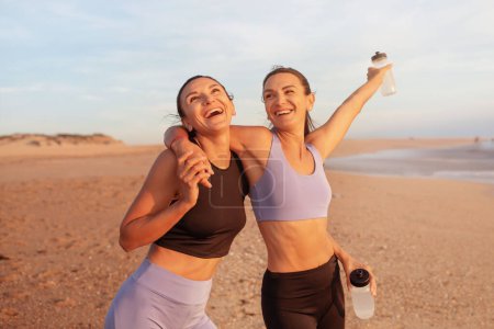 Photo for Satisfied cheerful young caucasian women hugs, enjoy morning workout, have fun with water bottles on sea beach. Vitality, sports, active lifestyle and body care, weight loss outdoor - Royalty Free Image