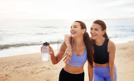 Téléchargez les photos : Rire joyeux jeunes femmes caucasiennes embrasser, avec des bouteilles d'eau, se reposer de l'entraînement, amusez-vous sur la plage de la mer. Sport, fitness ensemble, lifestyle, pilates et soins du corps, outdoor - en image libre de droit