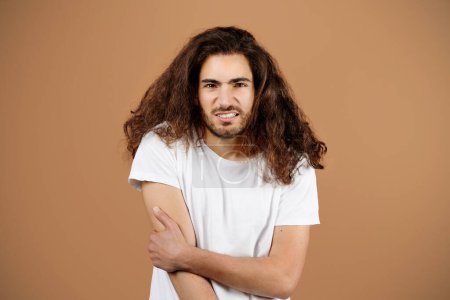 Téléchargez les photos : Jeune homme hispanique peu confiant aux cheveux longs posant main touchante souffrant de doutes et de préoccupations, se sentant incertain et mal à l'aise debout sur fond de studio beige - en image libre de droit