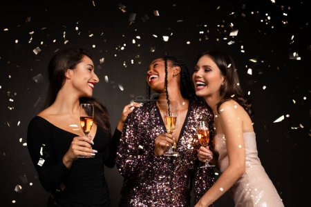 Foto de Tres hermosas damas jóvenes felices en bonitos vestidos posando en confeti sobre fondo negro, novias multirraciales celebrando el Año Nuevo juntas, bebiendo champán, riendo, tienen fiesta de Navidad - Imagen libre de derechos