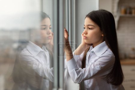 Foto de Lucha Emocional Juvenil. Lonely Preteen Girl Touching Window Glass, Gazing Out with Pensive Unhappy Expression at Home. Problemas de bienestar mental infantil, depresión infantil y soledad. Espacio libre - Imagen libre de derechos