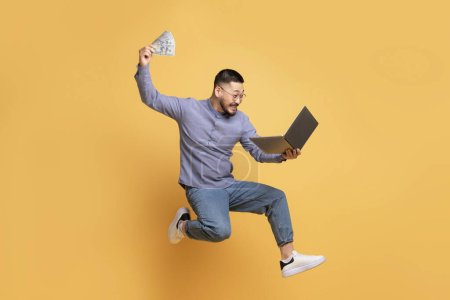 Big Profit. Excited Young Asian Man Jumping With Laptop And Dollar Cash In Hands On Yellow Background, Joyful Happy Millennial Guy Celebrating Successful Investment Or Online Lottery Win, Copy Space