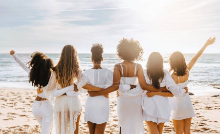Photo for Back view of multiracial ladies in white dresses posing on beach and looking at ocean, having maiden evening, hen party outdoors. Group of women embracing - Royalty Free Image