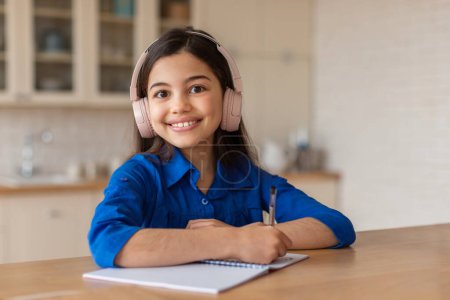 Foto de Estudio en casa. Happy School Girl se involucró en el aprendizaje, escribiendo en su cuaderno con auriculares inalámbricos y sonriendo a la cámara, posando sentada en la mesa interior. Ocio Educativo, Tareas - Imagen libre de derechos