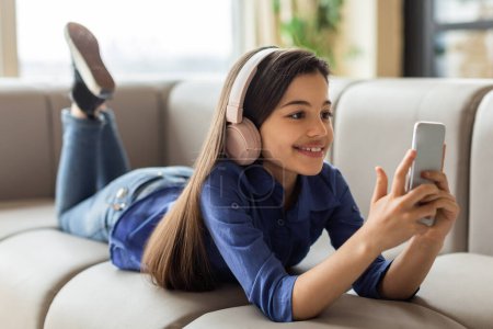 Foto de Diversión digital. Sonriente adolescente usando teléfono móvil usando auriculares inalámbricos, disfrutando de la música y viendo videos en línea, tumbado relajándose en el sofá en el interior del hogar. Niño y Gadgets - Imagen libre de derechos