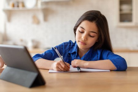 Foto de Educación remota. Joven niña preadolescente que estudia en la tableta digital sentada en el escritorio y escribiendo en su cuaderno en casa. Colegiala viendo curso en línea, conferencias y tomando notas - Imagen libre de derechos