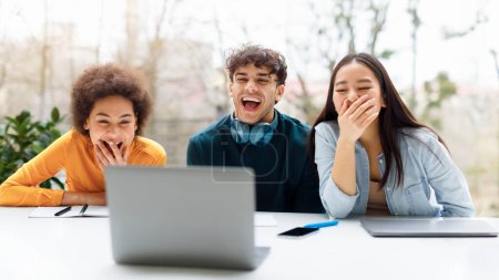 Téléchargez les photos : Heureux amis étudiants diversifiés regardant du contenu drôle sur ordinateur portable ensemble, assis dans un espace de coworking ou un public universitaire moderne et riant, panorama - en image libre de droit