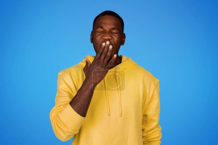 Téléchargez les photos : Fatigué millénariste homme afro-américain en sweat-shirt couvre sa bouche avec la main et bâillements, besoin de repos, isolé sur fond de studio bleu. Surmenage, sommeil, émotions humaines et ennui - en image libre de droit