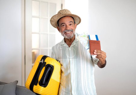 Foto de Feliz Globetrotter. Smiling Latin Mature Traveler Man Oferta turística de vacaciones publicitarias, Posando con maleta de viaje amarilla y boletos de tarjeta de embarque en casa, Usando sombrero de verano - Imagen libre de derechos