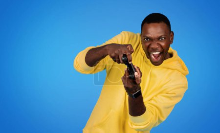 Photo for Happy excited millennial african american guy gamer in sweatshirt with open mouth and joystick playing online game, isolated on blue studio background. Ad and offer, win, fun and entertainment - Royalty Free Image