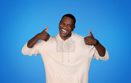 Téléchargez les photos : Joyeux millénium homme afro-américain en sweat-shirt montrer pouces vers le haut geste avec les mains, isolé sur fond bleu studio. Fun, annonce et offre, vente, recommandation et approbation - en image libre de droit