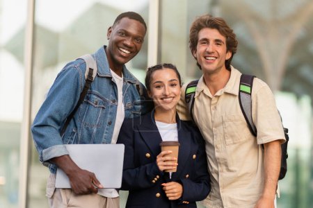 Photo for Happy cheerful young lady and two guys students with laptop in hands embracing and smiling outdoor, multiracial friends having fun after school, drink takeaway coffee, walk by city, copy space - Royalty Free Image