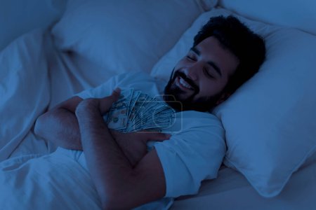 Foto de Hombre indio sonriente hablando con dólares en efectivo en las manos en la noche, calma relajado oriental masculino soñando con la riqueza y ser rico, sosteniendo billetes de dinero mientras duerme en el dormitorio oscuro, primer plano - Imagen libre de derechos