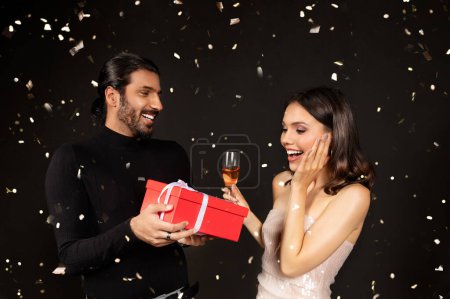 Photo for Amazed pretty young lady wearing elegant dress and nice makeup getting xmas present from her boyfriend, multiracial couple celebrating new year 2024 on black background, drinking champagne - Royalty Free Image