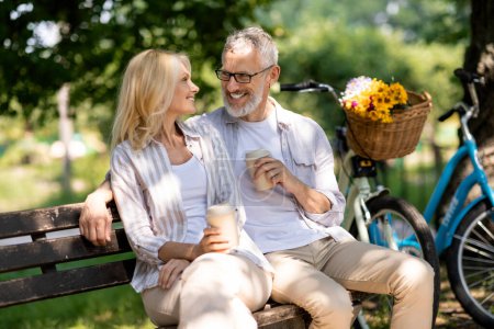 Téléchargez les photos : Couple d'âge mûr romantique avec café relaxant sur le banc dans le parc d'été, heureux homme et femme plus âgés embrassant et appréciant boisson à emporter, aimant les conjoints aînés bavarder et se regardant - en image libre de droit