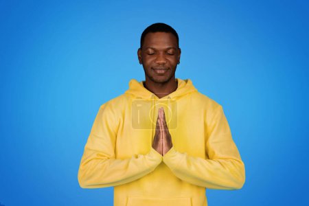 Photo for Smiling millennial african american guy prayer in hoodie with eyes closed making pray sign, isolated on blue studio background. Request, hope and faith, wish come true, dream - Royalty Free Image