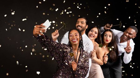 Téléchargez les photos : Heureux amis multiraciaux hommes et femmes attrayants dans de belles tenues nettoyer le Nouvel An ensemble, prendre selfie sur smartphone parmi les confettis tombant sur fond noir, tenant photomaton pops - en image libre de droit