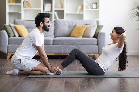 Téléchargez les photos : Formations domestiques. Couple indien athlétique s'exerçant ensemble à la maison, travaillant sur les muscles abdominaux sur tapis de fitness dans le salon, homme tenant les jambes de sa petite amie tout en faisant des exercices pour le noyau - en image libre de droit