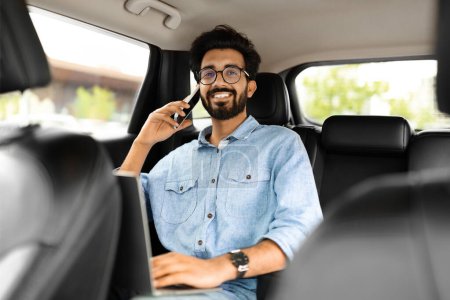 Photo for Always available. Handsome young eastern businessman in casual wearing eyeglasses working on his laptop and talking on the phone while sitting in the car back seat, copy space - Royalty Free Image