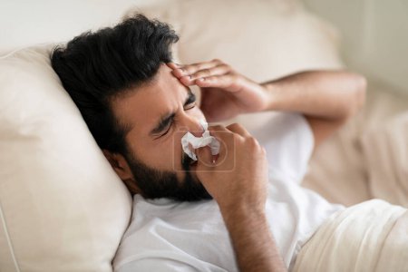 Téléchargez les photos : Jeune Indien malade qui souffle du nez dans des tissus de papier alors qu'il est allongé au lit à la maison, un homme de l'Est souffrant de migraine, de grippe saisonnière ou de rhume, ayant une rhinite ou une réaction allergique, gros plan - en image libre de droit