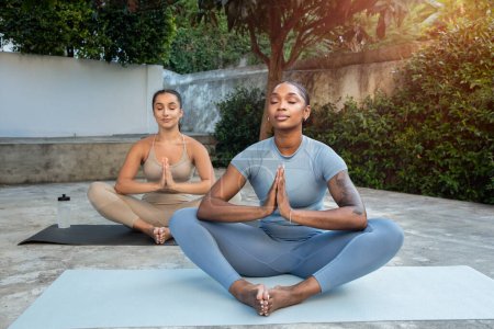 Photo for Positive calm millennial black and arab women do breathing exercises in lotus position, meditation, practice yoga together on mat, enjoy workout, outdoor. Health care, fit at morning - Royalty Free Image