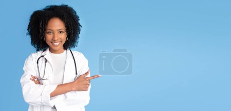 Téléchargez les photos : Femme médecin millénariste noire positive en manteau blanc pointe du doigt sur l'espace de copie, isolée sur fond de studio bleu. Conseil, recommandation, travail professionnel, médecine et soins de santé - en image libre de droit