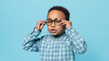 Eyesight. Portrait of little black boy wearing eyeglasses looking at camera, standing on blue studio background. Kids eyes health and sight correction concept