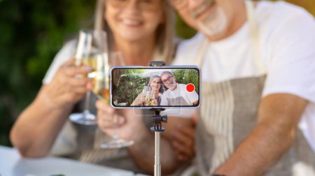 Photo for Smiling old caucasian husband and wife cheers glasses of wine in garden, shoot video on smartphone screen outdoor. Romantic date at home, dinner and relationship, holiday celebration with gadget - Royalty Free Image