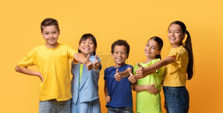 Photo for Multiethnic group of kids friends posing on yellow studio background, showing thumb up and smiling at camera. Happy children boys and girls recommending something nice, enjoying time together, banner - Royalty Free Image