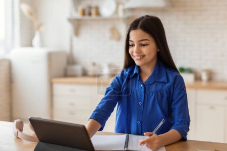 Photo for E-Learning Leisure. Happy school learner girl using digital tablet and taking notes at kitchen interior, studying online from home. Preteen schoolgirl watching webinar on computer, doing homework - Royalty Free Image