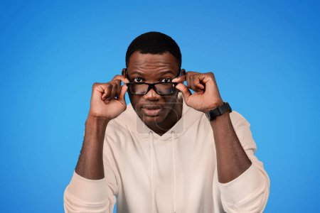 Foto de Interesado curioso millennial afroamericano chico en sudadera se quita las gafas, mira a la cámara, comprobación, aislado en el fondo del estudio azul. Pobre vista, anuncio y oferta - Imagen libre de derechos