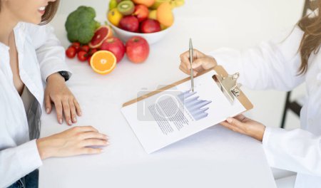 Photo for Adult caucasian nutritionist doctor in white coat shows weight loss plan to young lady patient in clinic office interior. Proper nutrition, recommendation and advice by professional, health care - Royalty Free Image