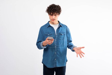 Photo for Strange Phone Message, Bad Mobile News. Confused curly guy holds cellphone and has problem with gadget and internet connection on smartphone, shrugging shoulders standing on white background - Royalty Free Image