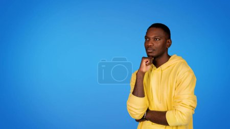Téléchargez les photos : Pensif homme afro-américain millénaire à capuche penser, en regardant l'espace de copie, isolé sur fond de studio bleu, panorama. Annonce et offre, vente, remue-méninges et choix, création d'idées, planification - en image libre de droit