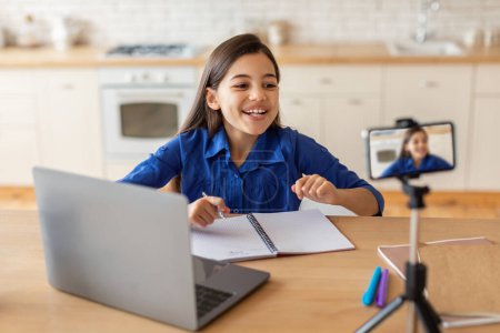 Photo for Modern E-Learning. Cheerful Arab Schoolgirl Having Online Lesson Via Video Call On Cellphone, Using Laptop And Taking Notes, Studying Remotely Watching Educational Lecture At Home Interior - Royalty Free Image