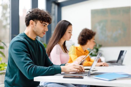 Téléchargez les photos : Étudiants multiraciaux masculins et féminins étudiant pour le test, faire des devoirs avec des gadgets, gars concentré à l'aide d'un ordinateur portable, assis à table dans un espace de coworking - en image libre de droit