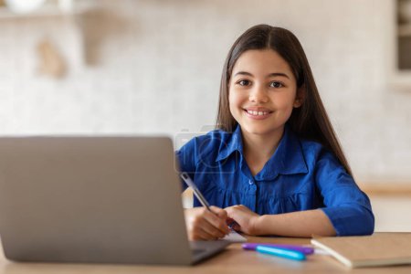 Photo for Distance Study. Teenage school girl posing at desk with laptop computer and writing in her notebook or diary, doing homework at PC indoors. Schoolgirl taking notes watching online lesson - Royalty Free Image
