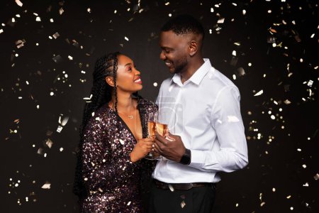 Foto de Amar a la joven pareja afroamericana en bonitos trajes celebrando el Año Nuevo sobre fondo negro entre confeti cayendo, abrazándose y mirándose, bebiendo champán, fiesta de Navidad - Imagen libre de derechos