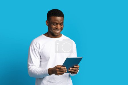 Foto de Chico negro joven guapo feliz en blanco usando la tableta digital moderna de la PC y sonriendo aislado en fondo azul, leyendo el ebook del blog de noticias en línea, desplazándose o websurf en Internet, espacio de la copia - Imagen libre de derechos