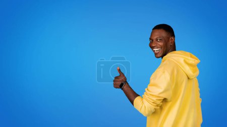Téléchargez les photos : Heureux homme afro-américain millénaire en sweat-shirt s'amuse, faisant geste pouce vers le haut, isolé sur fond de studio bleu, panorama. Annonce et offre, vente, recommandation et approbation de l'étude - en image libre de droit