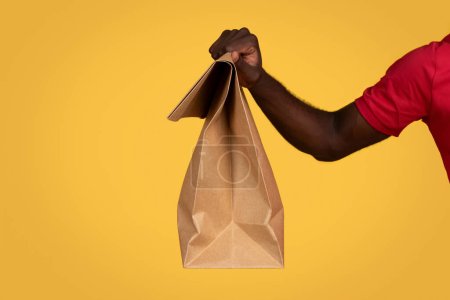 Photo for Hand of millennial african american guy courier hold paper bag with food takeaway, isolated on orange studio background. Fast delivery at home, work and service, ad and offer, cropped - Royalty Free Image