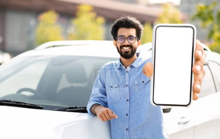 Photo for Happy young indian man standing by auto, showing smartphone with empty screen, showing newest mobile application for navigation, mockup. Car leasing, automobile, driving and modern technologies - Royalty Free Image