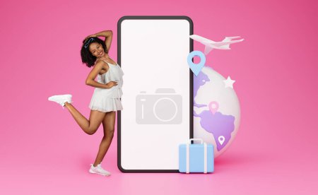 Téléchargez les photos : Jolie jeune femme afro-américaine insouciante et heureuse en tenue d'été choisissant une tournée de voyage en ligne, danse à côté d'un énorme smartphone avec écran blanc, valise et globe 3D, fond rose, collage - en image libre de droit