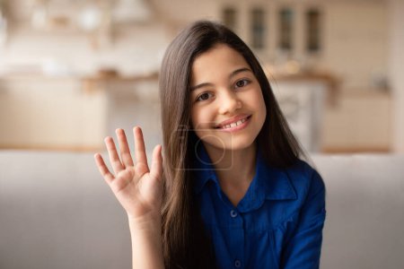 Foto de Hola. Hola. Retrato de niña preadolescente árabe alegre con el pelo largo agitando la mano en la cámara, gesto hola sentado en el interior de la casa moderna, posando con una linda sonrisa en la cocina en el interior - Imagen libre de derechos