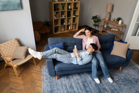 Foto de Pareja joven japonesa relajada usando Smartphone Descansando en el sofá en la sala de estar moderna en casa, tiro de ángulo alto. Los cónyuges asiáticos envían mensajes de texto a través de aplicaciones móviles y comparten contenido de redes sociales juntos - Imagen libre de derechos