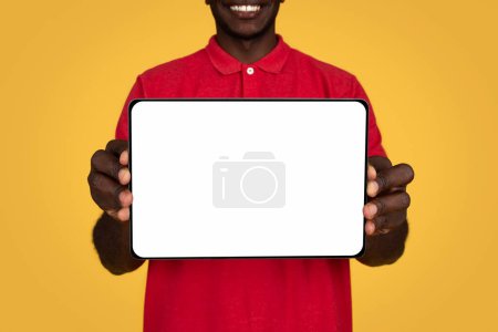 Foto de Positivo millennial afroamericano chico mensajero en uniforme mostrar tableta con pantalla en blanco, aislado en fondo de estudio naranja. Sitio web, aplicación, gadget para la entrega, trabajo y buen servicio, anuncio, oferta - Imagen libre de derechos