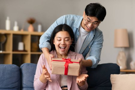 Photo for Loving Korean Boyfriend Giving Gift Box To Girlfriend, Surprising Her On Birthday Holiday Sitting On Couch At Home. Family Celebrating Special Date, Anniversary. Presents And Celebration Concept - Royalty Free Image